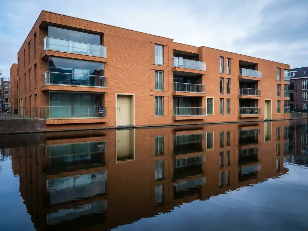 Modern appartementengebouw met glazen balkons weerspiegeld in Amsterdamse gracht, te koop bord bij ingang